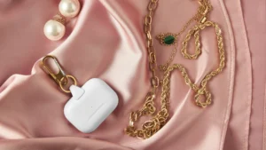 Gold chains, a green pendant necklace, pearl earrings, and a white wireless earbud case with a gold keychain, artfully arranged on pink silk fabric.