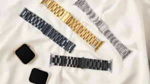 Set of four luxury metal Apple Watch bands in black, gold, silver, and blue, with two Apple Watches on a soft cream background.