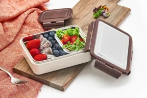 Plastic compartment lunch box by Pratap Plastics with strawberries, dragon fruit, blueberries, lettuce, cherry tomatoes, and microgreens, displayed on a wooden board with a fork and napkin.