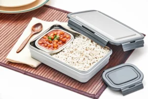 Stainless steel lunch box by Pratap Plastics with compartments of white rice and chickpea curry, placed on a placemat with wooden spoon and napkin.