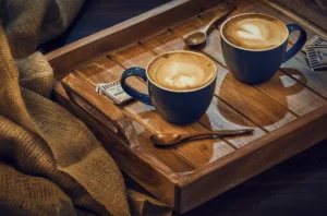 Two blue mugs of latte art coffee with heart designs on a wooden tray with wooden spoons and sugar packets, surrounded by burlap fabric.
