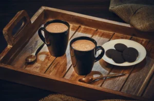 Two black mugs of hot chocolate on a wooden tray with two spoons, beside a white plate of dark chocolate cookies.
