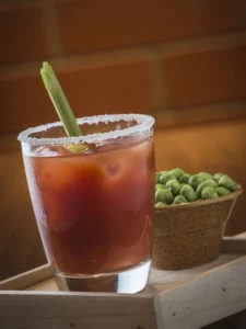 Glass of Bloody Mary with ice, salted rim, and celery stick garnish, next to a bowl of green peas, on a wooden tray.