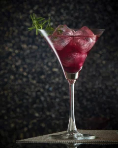 Deep red berry martini cocktail with ice cubes in a martini glass, garnished with a sprig of fresh rosemary against a dark, festive background.