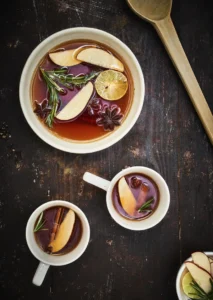 Overhead view of mulled cider with apple slices, citrus, rosemary, and star anise in a bowl and mugs on a rustic wooden table.