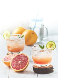 Two glasses of grapefruit and lime cocktail over ice, garnished with mint and lime slices, surrounded by halved grapefruit, whole grapefruit, and a glass jar of water on a white wooden surface.