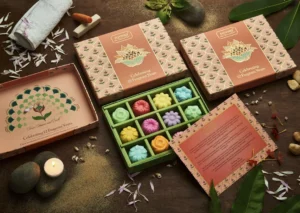 Assorted colorful flower-shaped soaps in a decorative Nyassa gift box with a lotus motif, surrounded by spa elements like candles, towel, and petals on a wooden table.