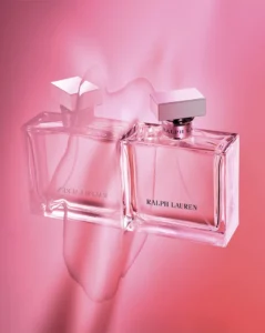 Ralph Lauren Romance perfume bottle with silver cap and minimalist clear glass, reflected on a pink surface against a soft pink background.