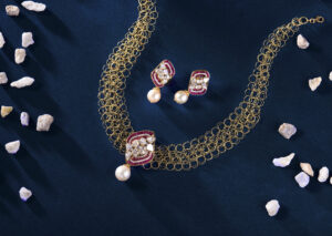 Handcrafted gold filigree necklace and matching earrings with pearls, ruby accents, and polki stones, displayed on a dark blue background with scattered white stones.