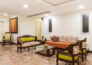 Traditional Indian living room with carved wooden sofas, floral upholstered cushions, a glass coffee table, marble floor, and warm wall art.