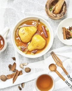 Bowl of poached pears in spiced syrup with whole cinnamon, star anise, and cardamom pods, surrounded by wooden utensils, tea cups, and loose spices on a white textured cloth.