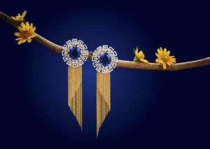 Pair of blue gemstone statement earrings with geometric clear crystals and long gold tassel chains displayed on a branch with yellow flowers against a deep blue background.