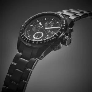 Black Fossil chronograph wristwatch with steel bracelet, tachymeter bezel, and multifunction subdials on a dark gradient background.