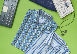 Two folded geometric-print shirts from hippieshippie, one with blue triangle motifs and another with blue dash patterns, laid on a green background alongside denim jeans, earphones, a paperback book, and a music controller.