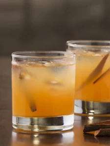 Glass of iced orange cocktail with cinnamon stick garnish, served on a wooden surface with another glass and extra cinnamon sticks nearby.