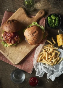 Overhead view of two bacon cheeseburgers with lettuce on a wooden board, served with French fries, steamed broccoli, grilled corn on the cob, ketchup, and a glass of water on a rustic table.