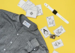 Flat lay of a grey linen shirt, illustrated elephant playing cards, white smart watch, and small electronic component on a bright yellow background.