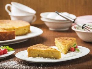 Two slices of golden semolina coconut cake on a white plate with grated coconut and berries, with the whole cake and kitchen bowls in the background.