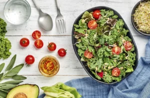 Overhead view of a fresh green salad with cherry tomatoes, mixed leafy greens, sprouts, avocado, water, herbs, and salad dressing on a white wooden background.