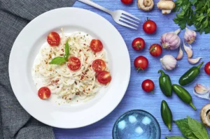 Overhead view of creamy white sauce spaghetti pasta garnished with cherry tomatoes, basil, and chili flakes, surrounded by fresh vegetables and herbs on a blue table.
