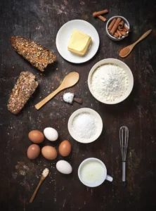 Overhead view of assorted baking ingredients: whole eggs, flour, butter, milk, shredded coconut, salt, cinnamon sticks, sliced multigrain bread, and kitchen tools on a dark rustic table.