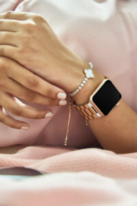 Close-up of a woman’s hand wearing a rose gold smartwatch and diamond bracelet on a light pink background