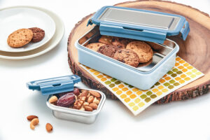 Pratap Plastic lunchbox set with stainless steel inner containers, filled with cookies, dry fruits, and nuts on a wooden background.