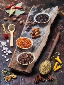 Assorted Indian spices including cinnamon, black pepper, cloves, coriander seeds, turmeric, star anise, nutmeg, chili flakes, cumin, saffron, bay leaves, and rock salt displayed on rustic wooden background.