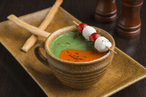 "Spinach and tomato soup served in a rustic brown cup, with a skewer of mozzarella balls and cherry tomatoes, paired with crisp breadsticks on a stoneware plate".