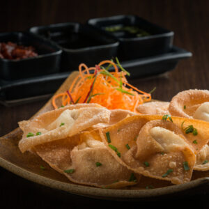 "Crispy fried wontons garnished with fresh chives, served on a brown plate with carrot salad and trio of dipping sauces in the background".