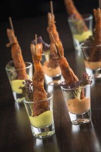 "Crispy fried prawns served upright in shot glasses with colorful dipping sauces and lettuce, styled on a dark surface".