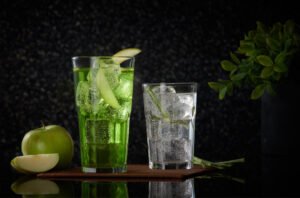 "Green apple soda mocktail and sparkling soda served in tall and short glasses over ice, garnished with green apple slices, with whole apple and plant decor".