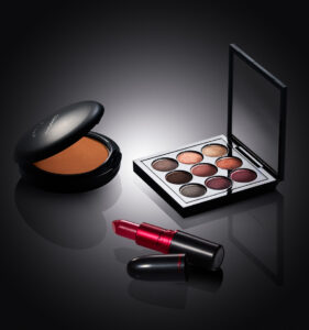 "Luxury MAC makeup set with pressed powder compact, nine-shade eyeshadow palette, and red lipstick displayed on a black reflective surface".