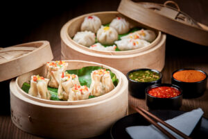 "Assorted dim sum including shumai and steamed dumplings, arranged on banana leaves in bamboo steamers, with three colorful dipping sauces and chopsticks".