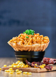 Crispy golden corn kernels served in a fried edible bowl, garnished with fresh herbs and surrounded by corn and dried chilies on a wooden surface