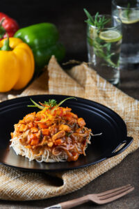 "Indo-Chinese vegetable noodles topped with bell peppers, carrots, and garnish served on a black plate, with fresh bell peppers and mocktail in the background".