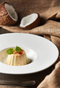 "Coconut panna cotta served on a white plate, garnished with fresh mint leaves and set against a rustic background with cracked coconuts".