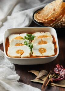 "Stuffed paneer slices served in tomato curry, garnished with coriander leaves and accompanied by crisp papad in a black bowl, with traditional Indian spices"