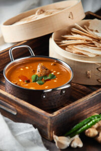 "Traditional Indian curry garnished with curry leaves and cream, served in a hammered copper bowl beside bamboo baskets filled with crispy papadum slices".