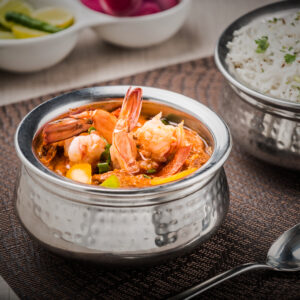 "Indian prawn curry served in a hammered silver bowl, garnished with fresh prawns, yellow bell peppers, and green beans, accompanied by a bowl of jeera rice".